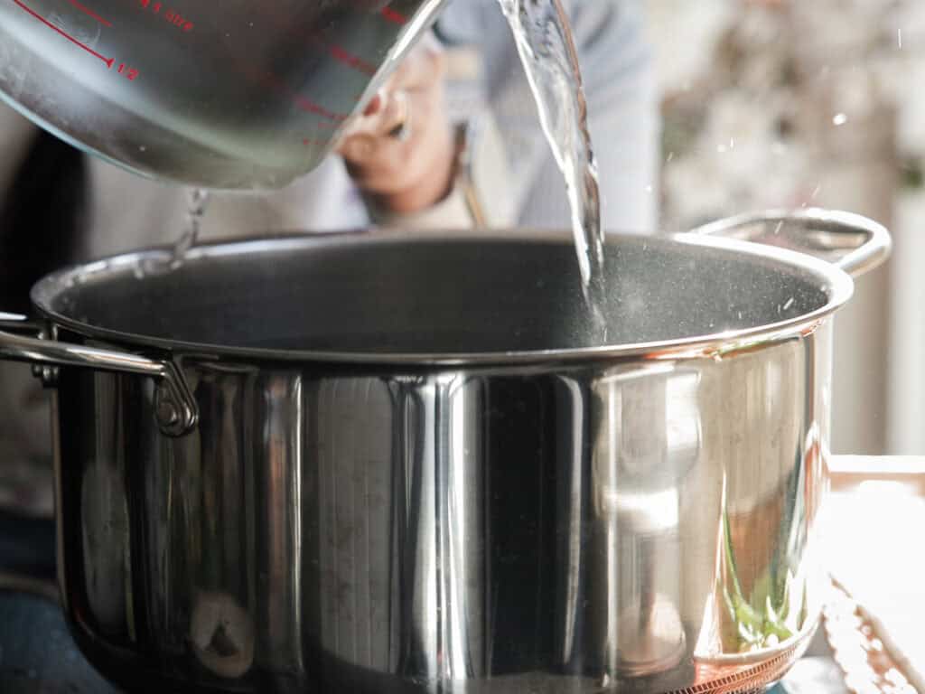 A person is pouring water from a large measuring cup into a shiny stainless steel pot on a stove. The setting appears to be a kitchen, with blurred background details.